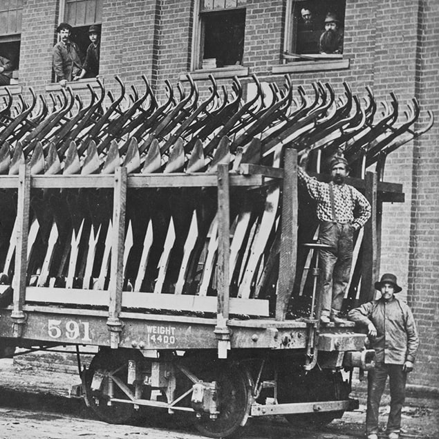 Foto uit 1882 met drie mannen die naast een Deere & Co treinwagon staan die volgeladen is met stalen ploegen voor verzending, en op de achtergrond fabrieksmedewerkers die uit het raam van het gebouw meekijken