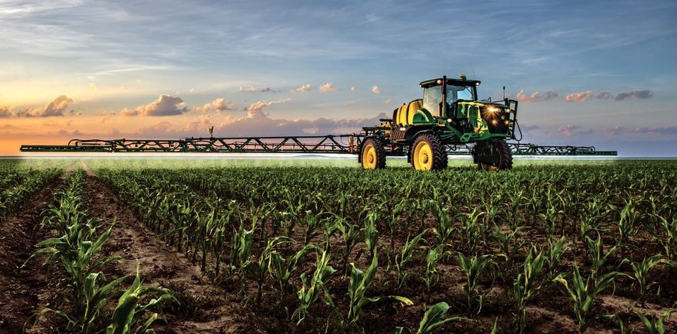 Pulvérisateur John Deere à l'œuvre dans un champ de maïs sur fond de coucher de soleil