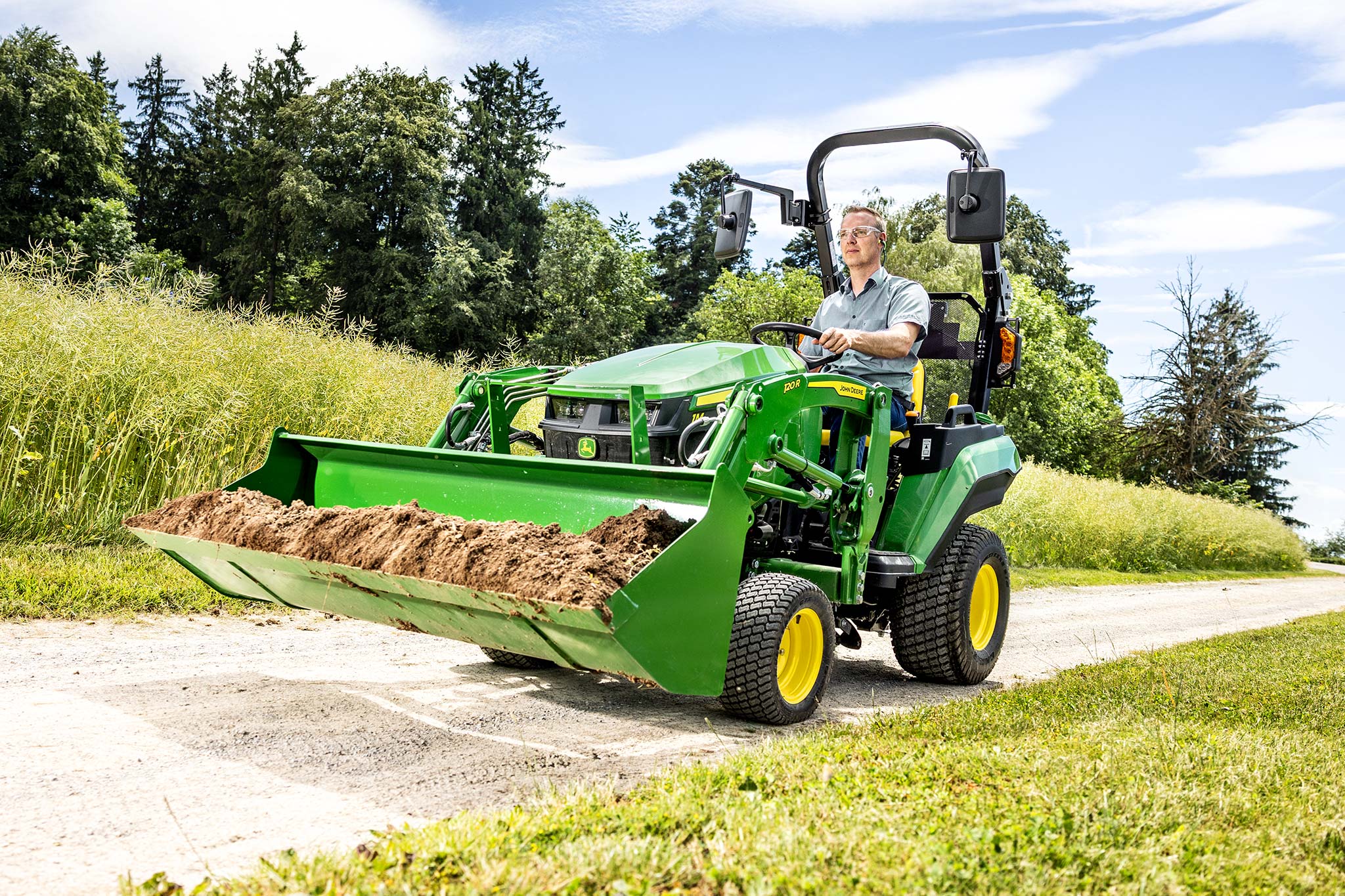 Un utilisateur au volant d’un tracteur compact John Deere transportant de la terre dans le godet de son chargeur frontal sur une route de campagne