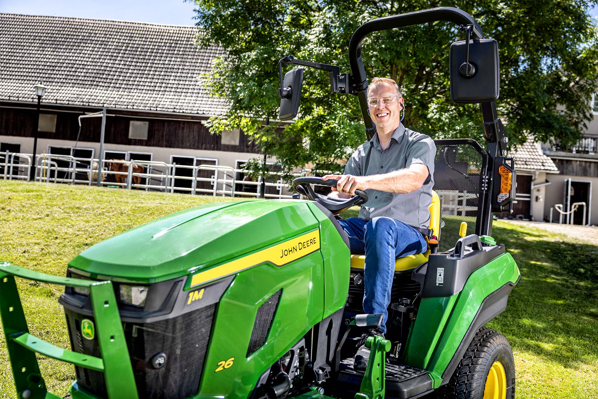 Un utilisateur au volant d’un tracteur compact John Deere à l’extérieur, à proximité de bâtiments agricoles et d’enclos clôturés pour le bétail