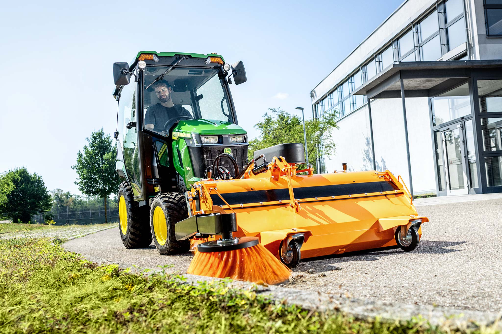 Tracteur compact John Deere équipé d’une cabine fermée et d’un balai rotatif monté à l’avant, en train de nettoyer une surface pavée