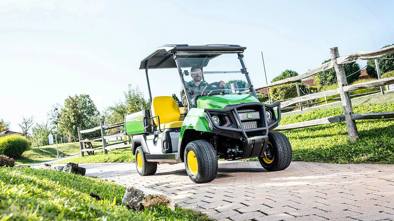 Véhicule utilitaire Gator&nbsp;GS John&nbsp;Deere vert roulant sur un chemin pavé à travers un espace paysager avec de l’herbe et des clôtures en bois.