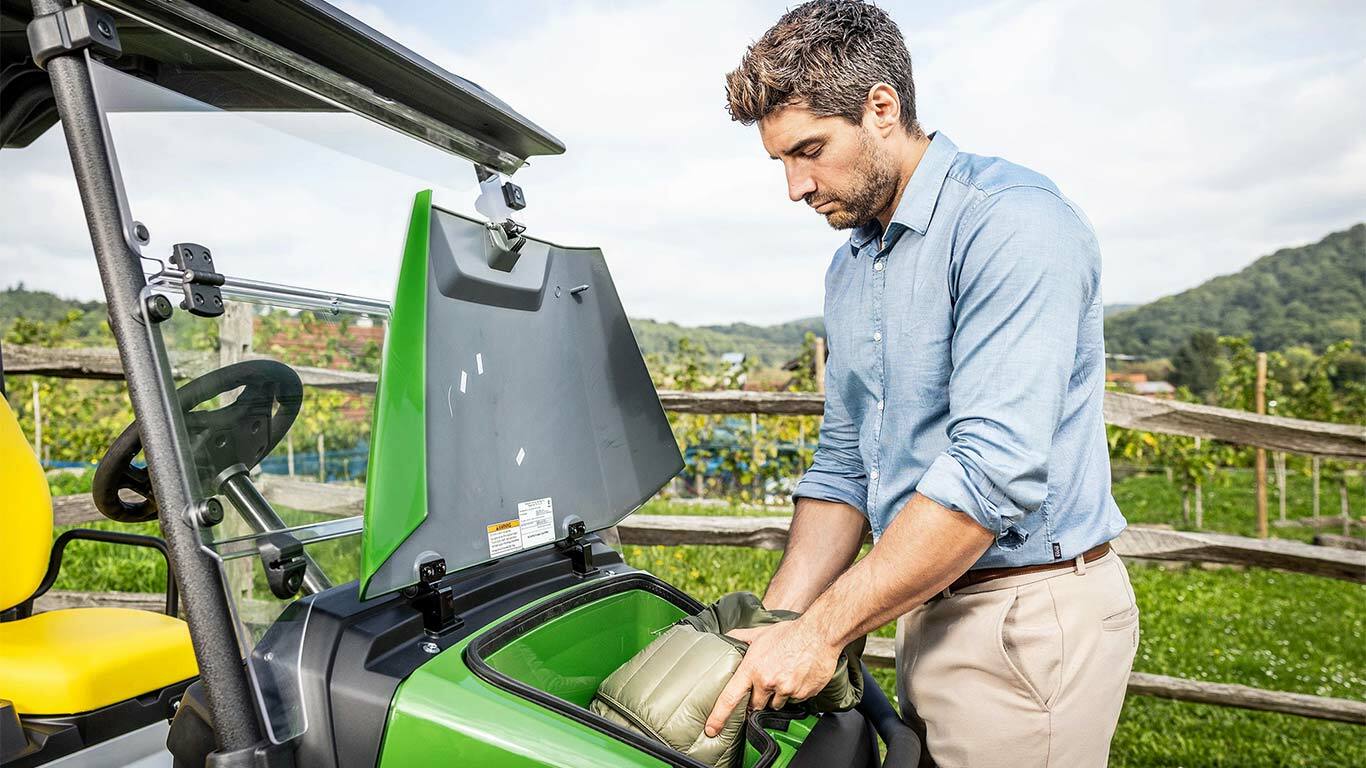 Une personne plaçant un sac dans le compartiment de rangement avant d’un Gator&nbsp;GS John&nbsp;Deere vert garé à l’extérieur près d’une clôture en bois et d’un vignoble.