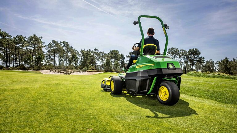 Une personne conduit une tondeuse de greens électrique John Deere 2775 E-Cut™ verte sur un parcours de golf bien entretenu, avec des arbres et un ciel partiellement nuageux en arrière-plan.