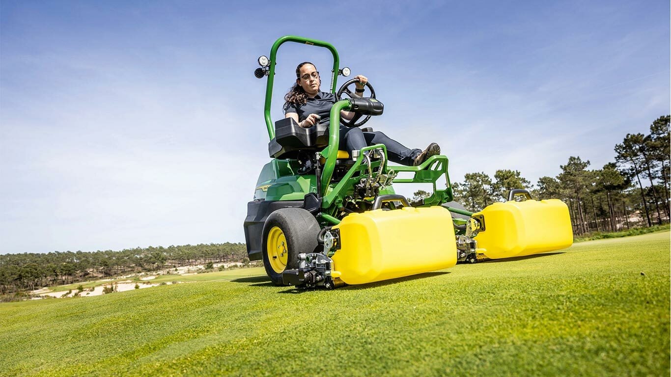 Une personne utilise une tondeuse électrique John Deere 2775 E-Cut™ vert et jaune avec des rouleaux avant couverts par un carter jaune sur un green de golf, avec des arbres et un ciel bleu clair en arrière-plan.