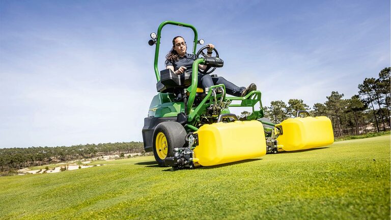 Une personne utilise une tondeuse électrique John Deere 2775 E-Cut™ vert et jaune avec des rouleaux avant couverts par un carter jaune sur un green de golf, avec des arbres et un ciel bleu clair en arrière-plan.