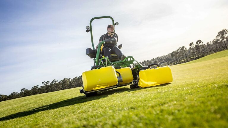Une personne utilise une tondeuse électrique John Deere 2775 E-Cut™ vert et jaune avec trois grands rouleaux avant recouverts de carters jaunes sur un green de golf, avec des arbres et un ciel partiellement nuageux en arrière-plan.