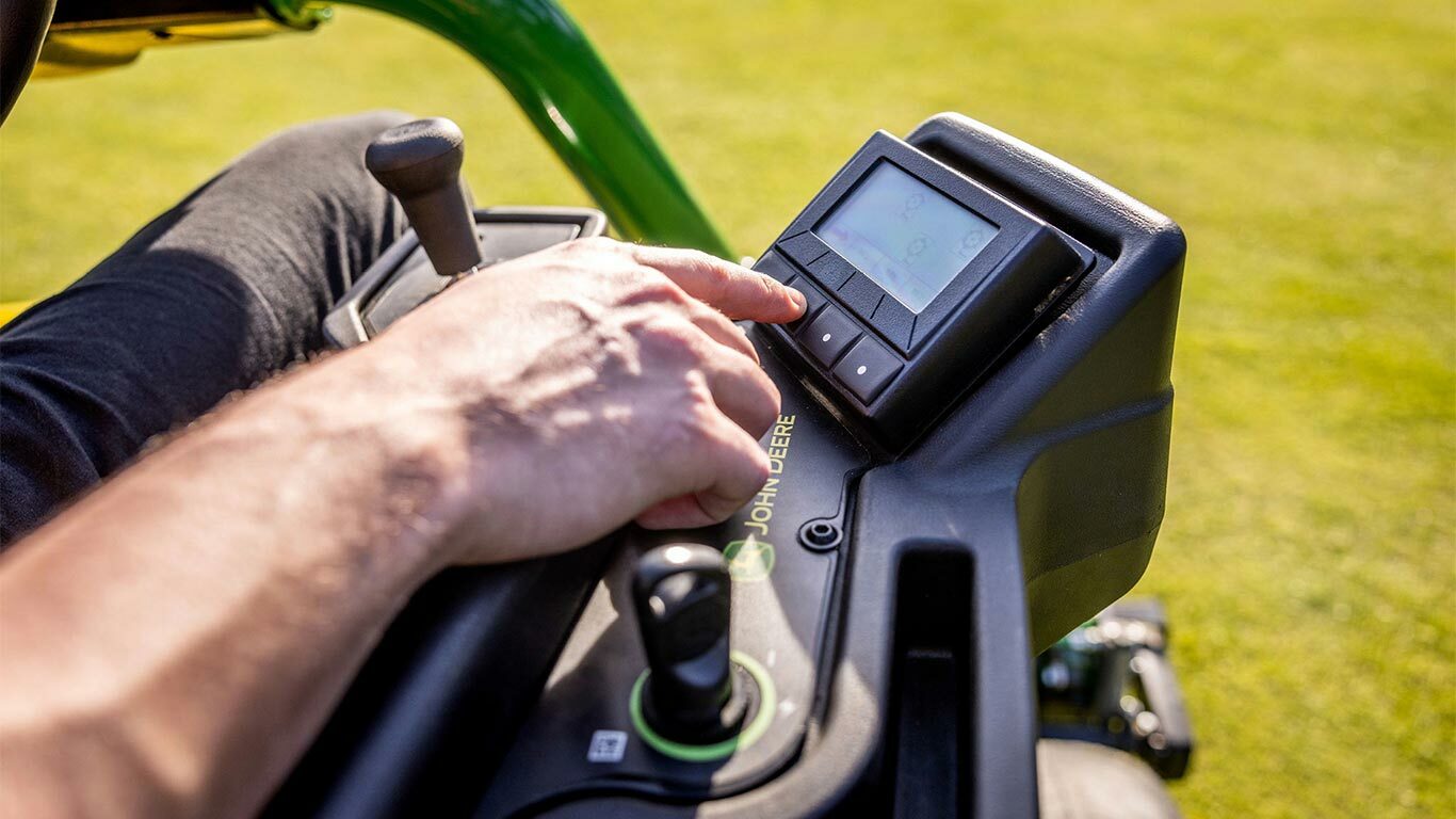 Gros plan de la main d’une personne utilisant le panneau TechControl d’une tondeuse électrique John Deere 2775 E-Cut™, appuyant sur les boutons d’une console numérique, avec un joystick et d’autres commandes visibles.