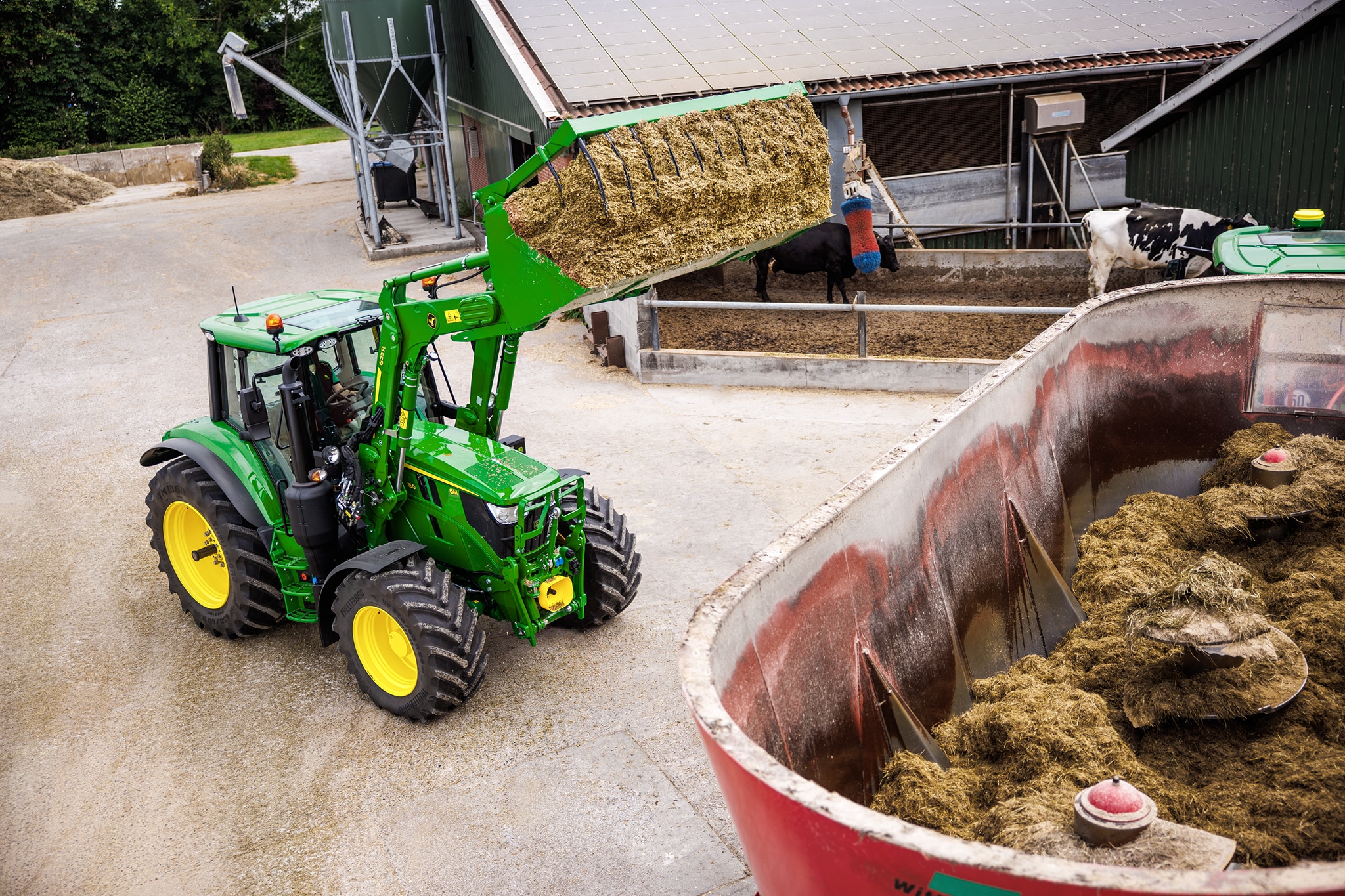 John Deere 6M Serie-tractor die met een voorlader kuilvoer in een voermengwagen laadt op een boerenerf.