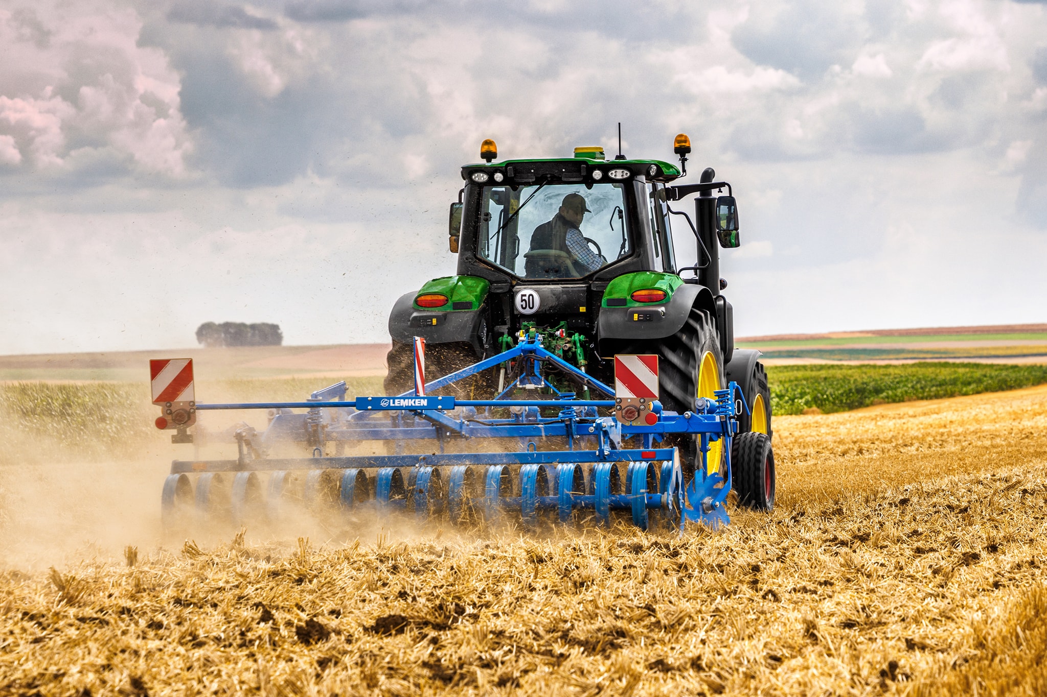 John Deere 6M Serie-tractor die een geoogst veld bewerkt met een schijveneg, achteraanzicht in stoffige omstandigheden.