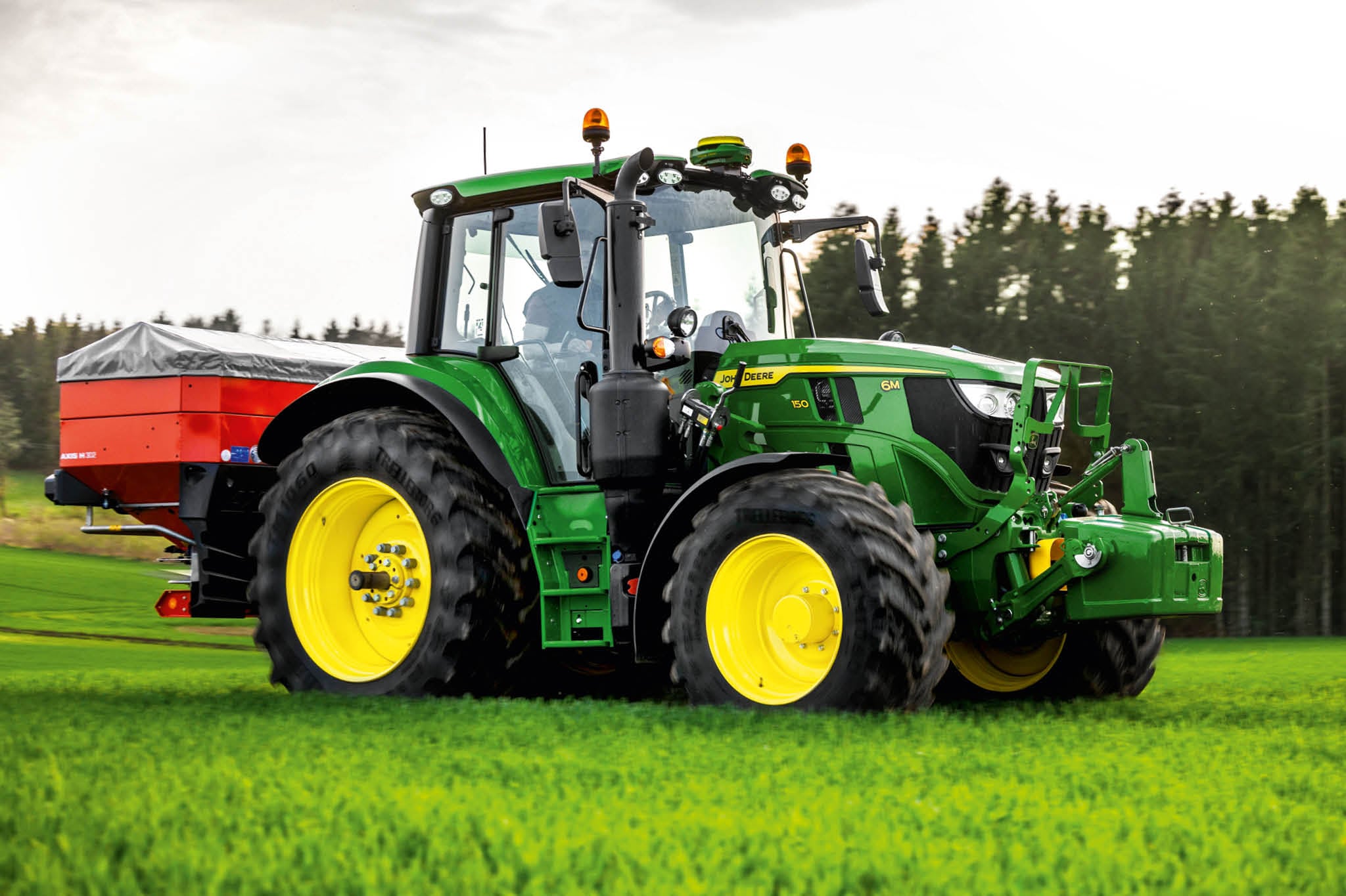 John Deere 6M serie tractor met achterop gemonteerde verdeler aan het werk op gras langs een bosrand.