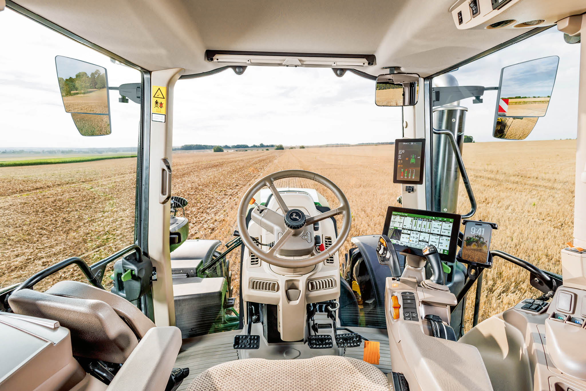 Intérieur de la cabine d’un tracteur John&nbsp;Deere, avec volant, consoles de commande numériques et vue sur le champ à travers le pare-brise pendant le travail.