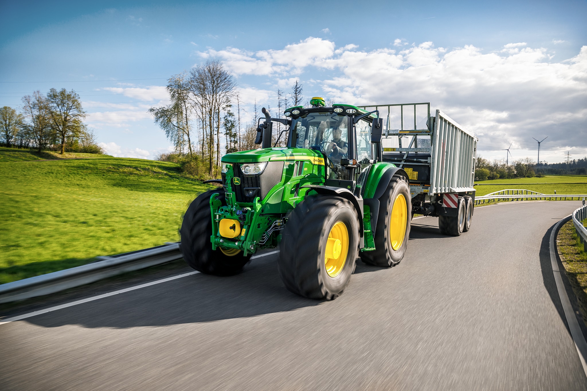 John Deere 6M Serie-tractor die een hoogwandige aanhanger vervoert op een verharde landelijke weg.