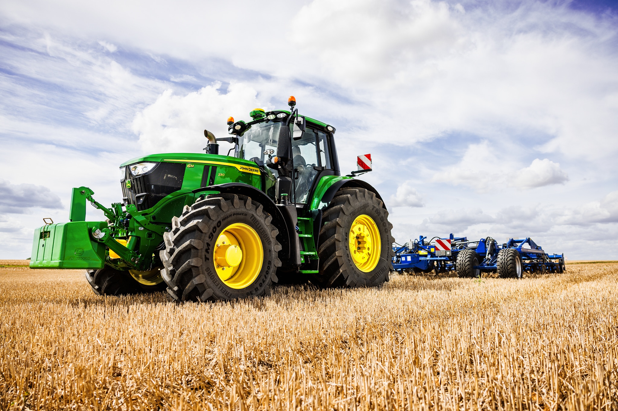 John Deere 6M Serie-tractor die een zwaar grondbewerkingswerktuig over een stoppelland trekt onder een bewolkte lucht.
