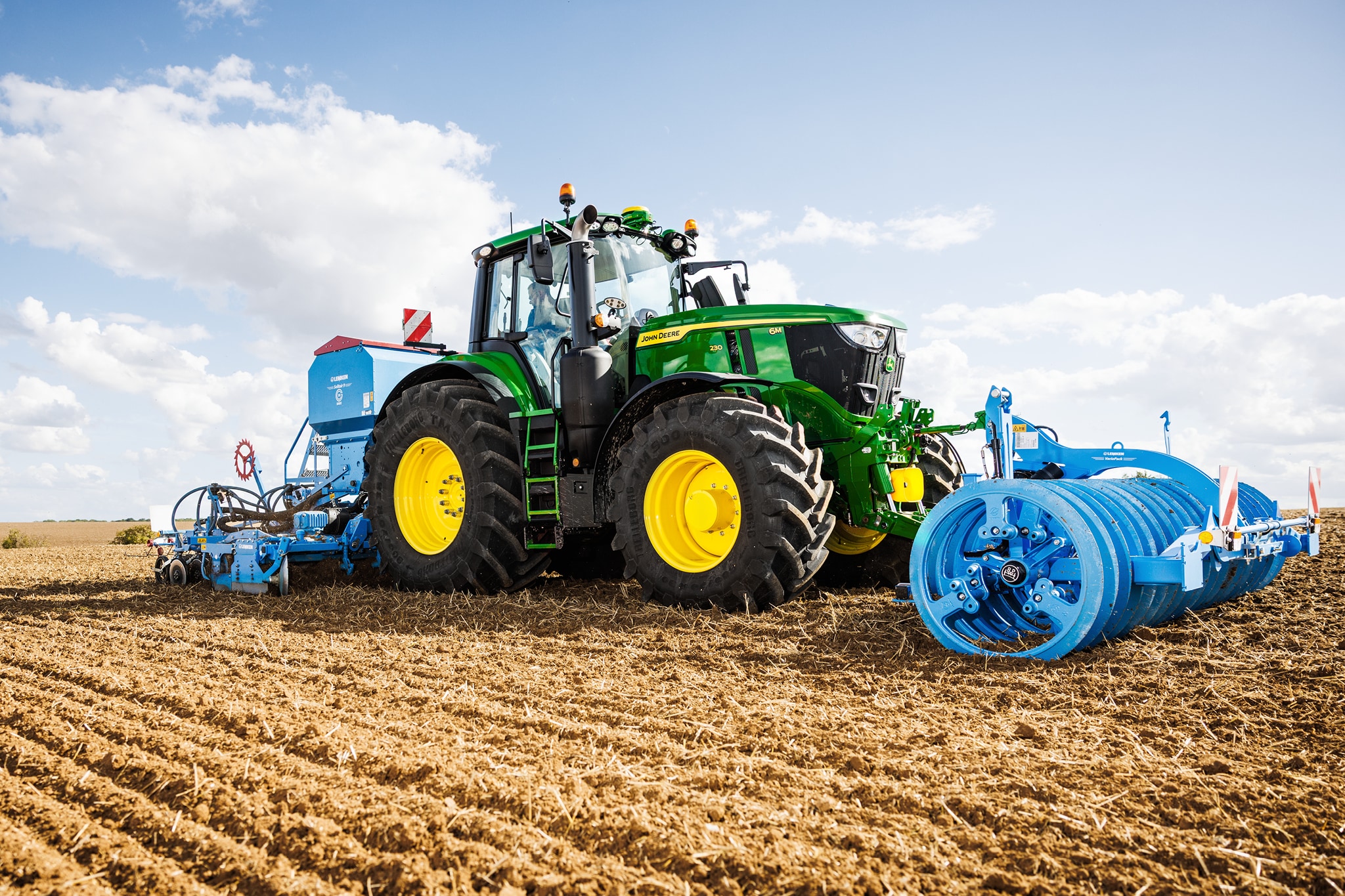 John Deere 6M Serie-tractor die een veld bewerkt met een grote blauwe rol.