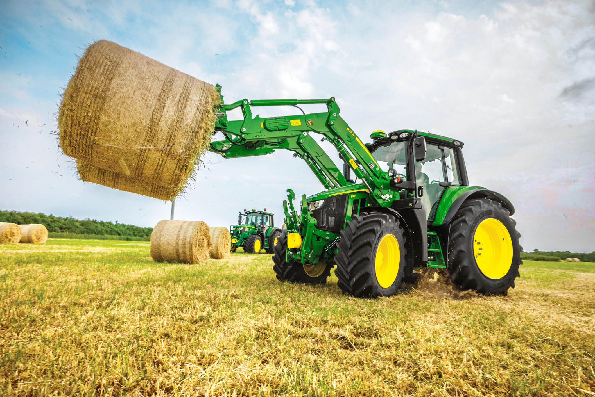 John Deere 6M Serie-tractor die een grote ronde hooibaal optilt met een voorlader op een open veld.