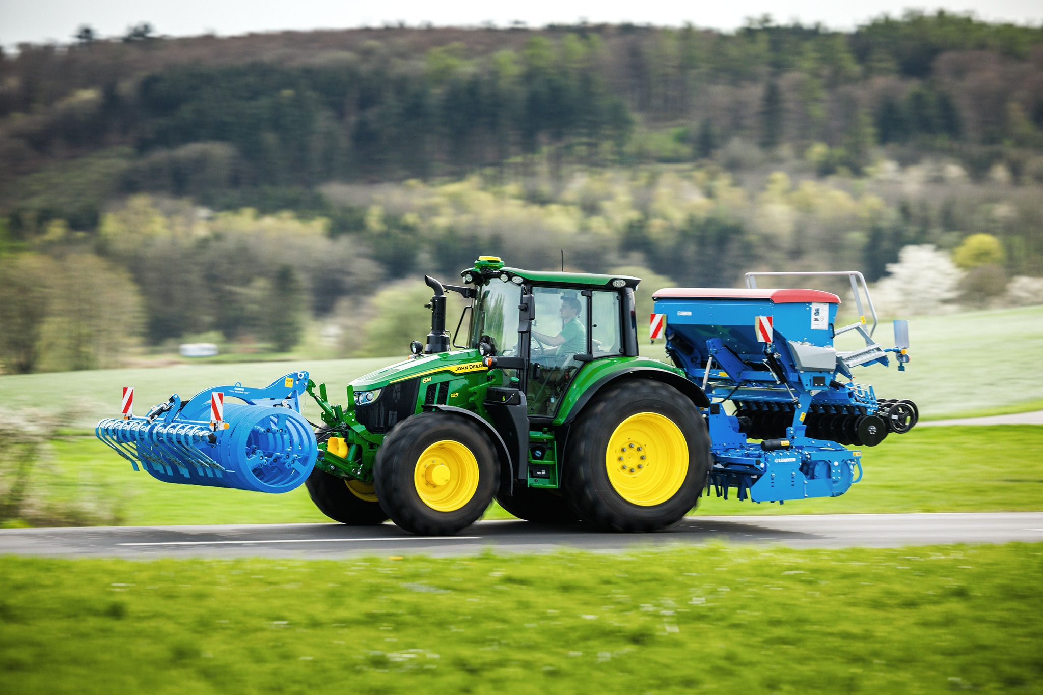John Deere 6M Serie-tractor die een voorop aangebouwd werktuig en zaaimachine achter transporteert over een landelijke weg.