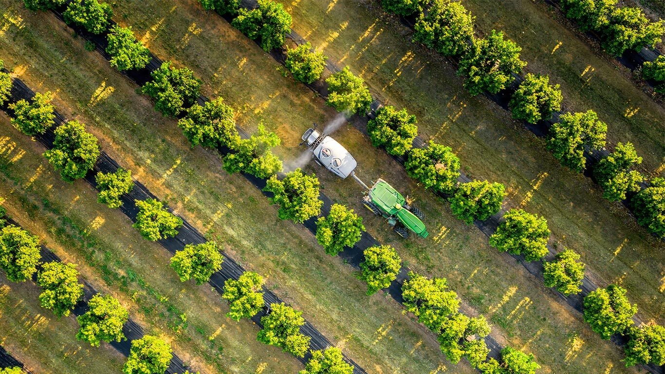 Vue aérienne d’un tracteur John Deere 5015EN vert et jaune remorquant un pulvérisateur à travers les rangs réguliers d’un verger.