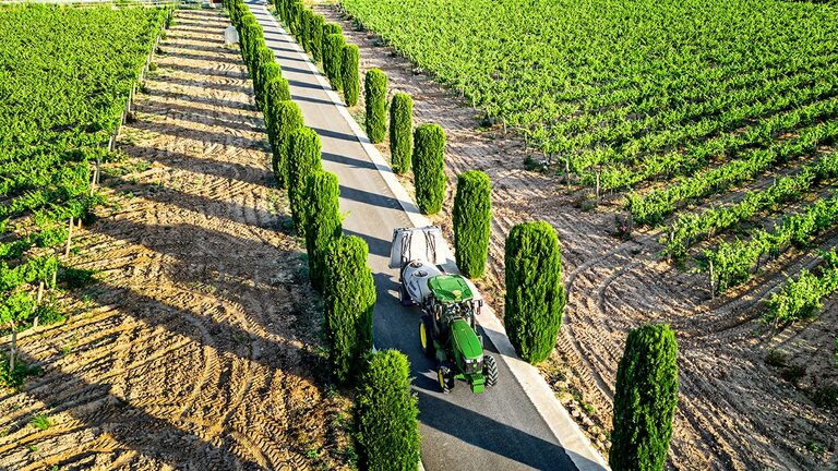 Tracteur John Deere 5075EN vert et jaune tirant un pulvérisateur le long d’un chemin bordé d’arbres entre les rangs de vignes, en plein soleil.