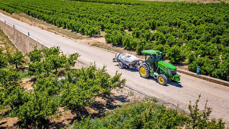 Tracteur John Deere 5090EN vert et jaune remorquant un pulvérisateur sur une route pavée entre de grands rangs de vergers sous un ciel bleu clair.