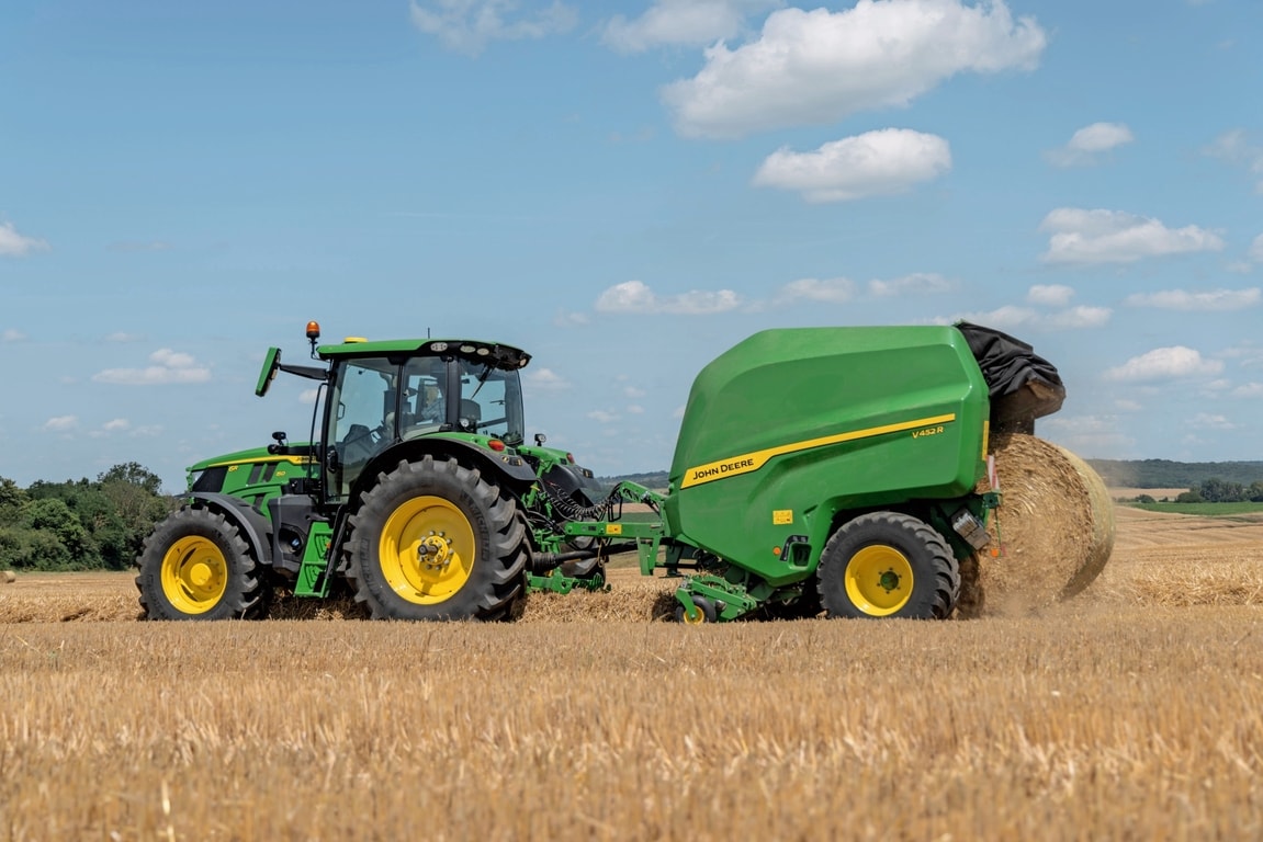 John Deere V452R dans un champ de paille tirée par un tracteur John Deere 6R 150 éjectant une balle