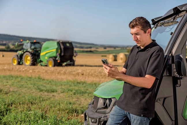 Personne vérifiant les données de la documentation des balles sur son téléphone portable avec une presse à balles VR John Deere en arrière-plan