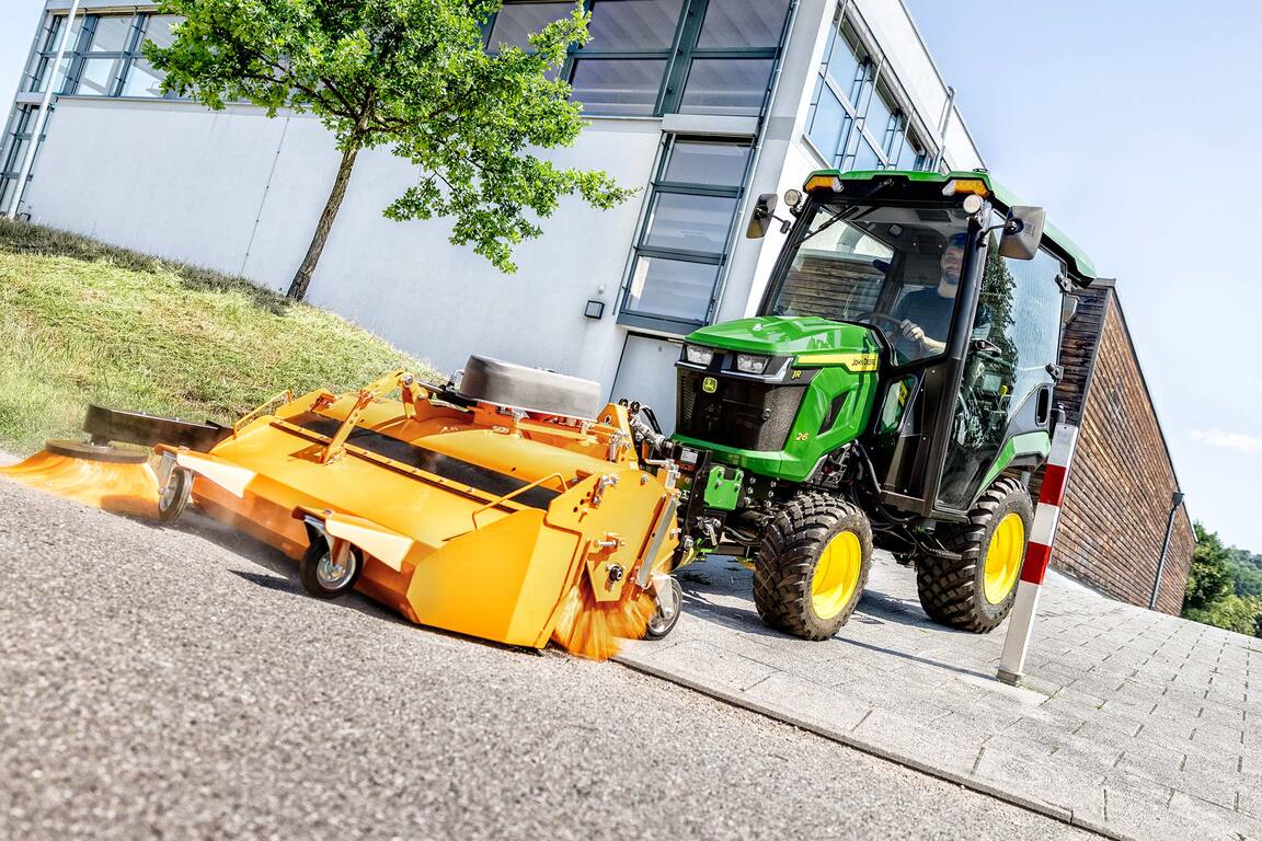 Tracteur compact John Deere équipé d’une cabine fermée et d’un balai rotatif à l’avant, balayant les feuilles sous les arbres à côté d’un bâtiment moderne