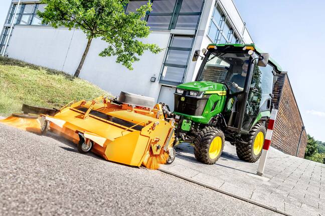 Tracteur compact John Deere équipé d’une cabine fermée et d’un balai rotatif à l’avant, balayant les feuilles sous les arbres à côté d’un bâtiment moderne