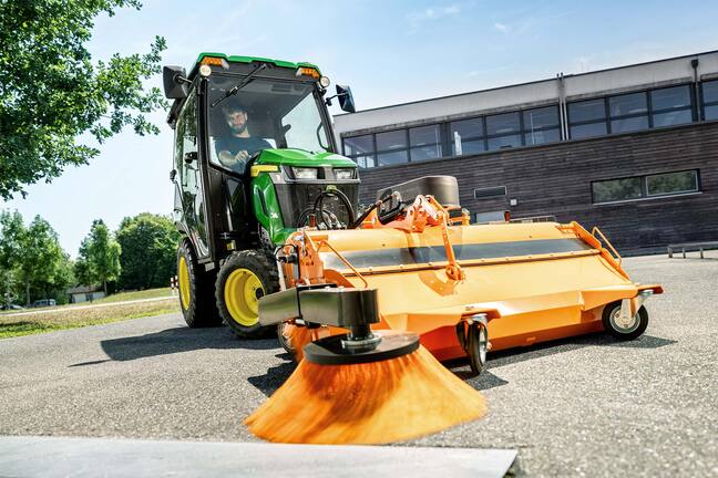 Tracteur compact John Deere équipé d’une cabine fermée et d’un balai rotatif avant, balayant les feuilles sous les arbres, dans une rue pavée