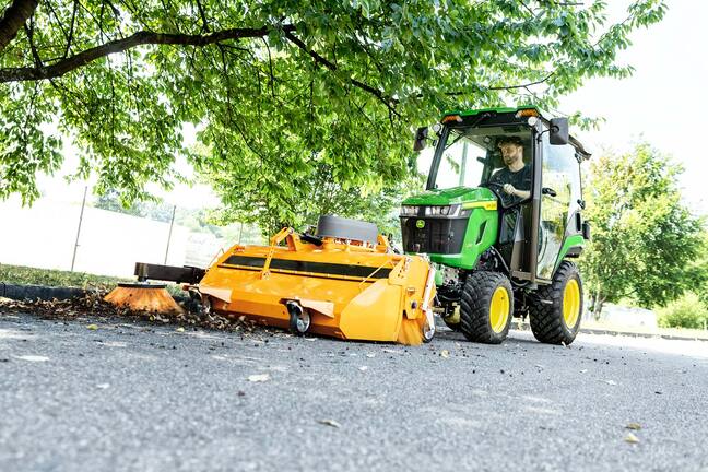 Tracteur compact John Deere équipé d’une cabine fermée et d’un balai rotatif avant, balayant les feuilles sous les arbres